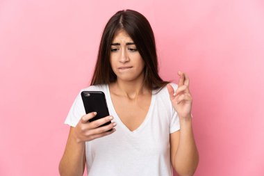 Young caucasian woman isolated on pink background using mobile phone with fingers crossing