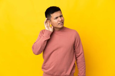 Genç Ekvadorlu adam sarı arka planda izole edilmiş bir şekilde elini kulağına koyarak bir şeyler dinliyor.