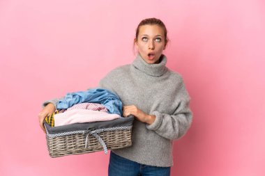 Pembe arka planda izole edilmiş çamaşır sepetini tutan genç bir kadın yukarı bakıyor ve şaşırmış bir ifadeyle