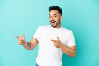 Mavi arka planda izole edilmiş genç beyaz adam şaşırmış ve işaret ediyor.