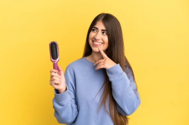 Mavi arka planda saç fırçası tutan genç beyaz kadın yukarı bakarken bir fikir düşünüyor.