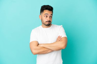 Kafkasyalı genç adam mavi arka planda izole edilmiş. Omuzlarını kaldırırken şüpheyle hareket ediyor.