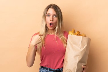 Elinde bej arka planda izole edilmiş bir çanta dolusu ekmek tutan sarışın bir kadın.