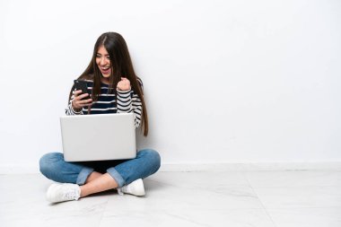 Young caucasian woman with a laptop sitting on the floor isolated on white background with phone in victory position