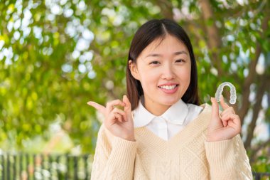 Dışarıda görünmez diş telleri tutan genç Çinli kadın şaşırdı ve parmağını yana çevirdi.