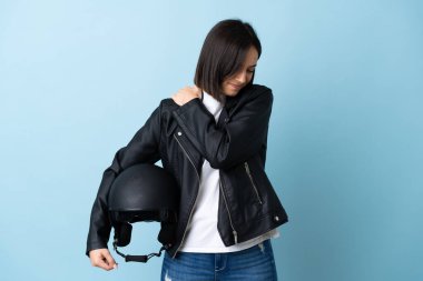 Mavi arka planda motosiklet kaskı tutan kadın, çabaladığı için omzunda ağrıdan muzdarip.