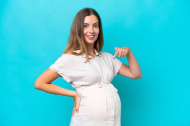 Mavi arka planda izole edilmiş genç beyaz kadın hamile ve karnını işaret ediyor.