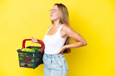 Beyaz bir kadın, elinde yiyecek dolu bir sepet tutuyor. Sarı arka planda izole edilmiş ve sırt ağrısı çekiyor.