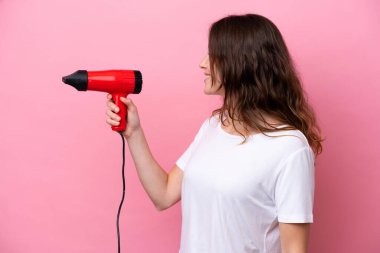 Kafkasyalı genç bir kadın elinde saç kurutma makinesiyle pembe arka planda mutlu bir ifadeyle izole edilmiş.
