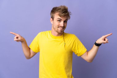 Mor arka planda izole edilmiş yakışıklı sarışın adam parmak uçlarını işaret ediyor ve mutlu.