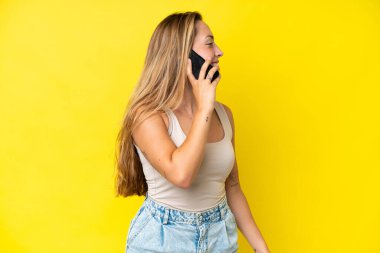 Sarı arka planda izole edilmiş genç beyaz bir kadın cep telefonuyla sohbet ediyor.