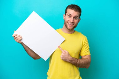 Young caucasian man isolated on blue background holding an empty placard with happy expression and pointing it