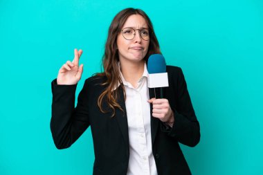 Genç TV sunucusu kadın, mavi arka planda izole edilmiş, parmaklarını çapraz yapıp en iyisini diliyor.