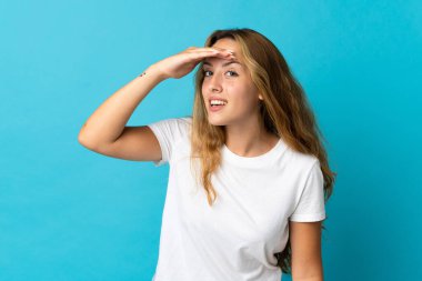 Mavi arka planda izole edilmiş genç sarışın bir kadın bir şeye bakmak için elini uzaklara bakıyor.