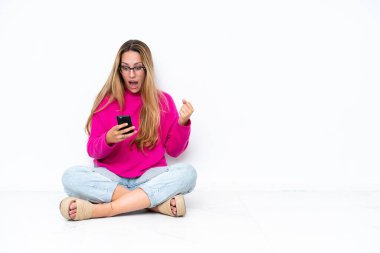 Beyaz arka planda izole bir şekilde yerde oturan beyaz bir kadın şaşırdı ve bir mesaj gönderiyor.