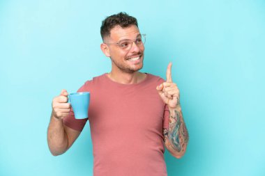 Kafkasyalı genç bir adam mavi arka planda izole edilmiş bir fincan kahve tutuyor. Çözümün farkına varmak için parmağını kaldırıyor.