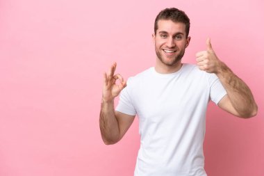 Pembe arka planda izole edilmiş genç beyaz bir adam tamam işareti ve baş parmak hareketi gösteriyor.