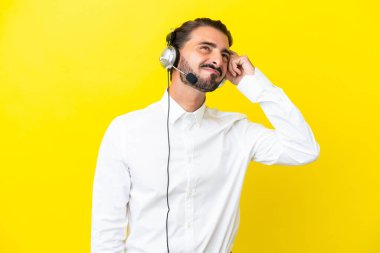 Tele pazarlamacı beyaz bir adam sarı arka planda izole edilmiş bir kulaklıkla çalışıyor. Şüpheleri var ve kafası karışmış bir yüz ifadesi var.