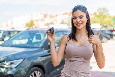 Dışarıda arabanın anahtarlarını tutan genç ve güzel bir kadın. Başparmakları havada çünkü iyi bir şey oldu.