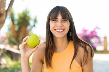 Açık havada elinde mutlu bir ifadeyle elma tutan genç bir kadın.
