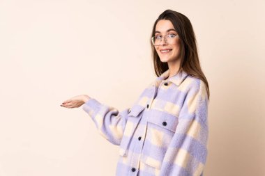 İzole edilmiş bej arka planda genç bir kadın. Davet edilmek için ellerini yana uzatıyor.