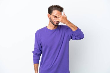 Beyaz arka planda izole edilmiş beyaz tenli genç bir adam. Gözleri elleriyle kaplıyor ve gülümsüyor.