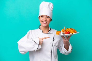 Kafkasyalı genç aşçı kadın elinde waffle tutuyor mavi arka planda izole edilmiş ve onu işaret ediyor.