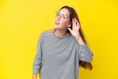 Sarı arka planda izole edilmiş genç beyaz bir kadın. Kulağına elini sürerek bir şeyler dinliyor.