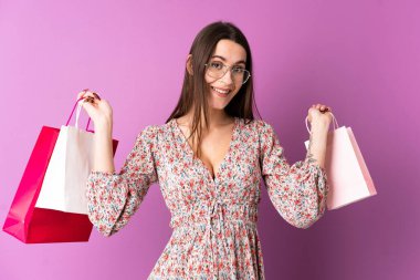 Soyutlanmış mor arka planda genç bir kadın alışveriş torbaları tutuyor ve gülümsüyor.