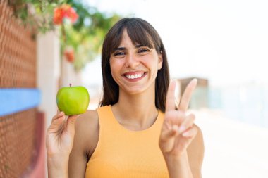 Dışarıda elmalı genç bir kadın gülümsüyor ve zafer işareti gösteriyor.