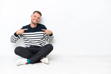 Yerde oturan genç adam beyaz arka planda kendini beğenmiş ve gururlu.