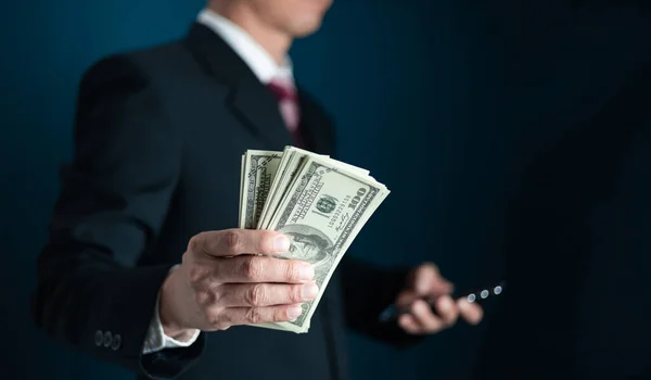 Takım elbiseli ve kravatlı iş adamı siyah arka planda elinde para tutuyor, Amerikan doları.