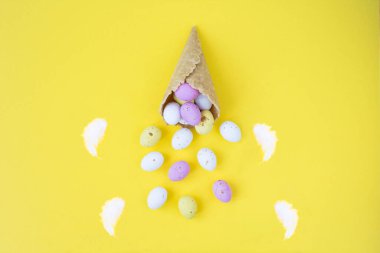 Renkli paskalya yumurtaları ve sarı arka planda beyaz tüylü dondurma külahı. Üst Manzara. Boşluğu kopyala.