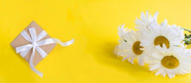 Gift box with tied white bow and white chamomile on the yellow background. Close-up. Copy space.