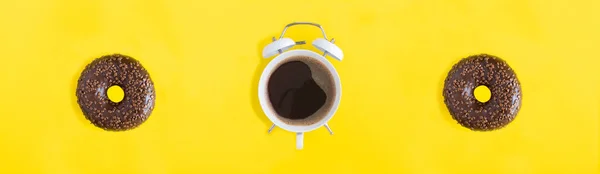 黑色咖啡在拨号盘上的白色闹钟和甜甜圈与巧克力釉面的黄色背景 顶部视图 — 图库照片