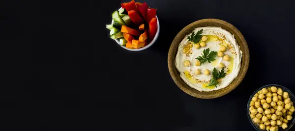 Hummus in the bowl and vegetable on the black background. Top view.  Copy space.