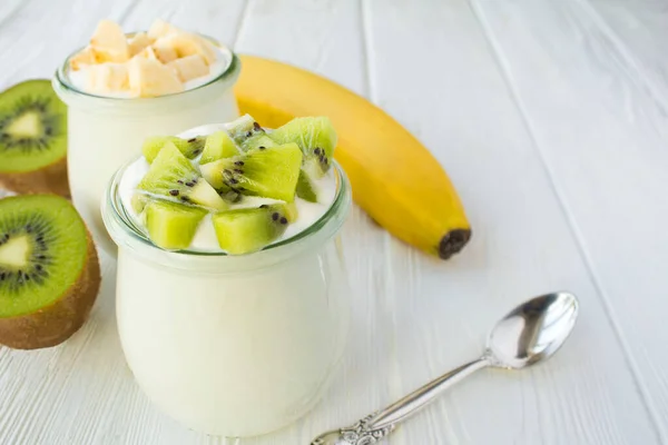 Natural milk yogurt with kiwi and banana in the glass jars on the white wooden background. Close-up.