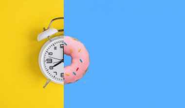 Beyaz alarm saati kolajı ve sarı ve mavi arka planda pembe jöleli donut. Uzayı kopyala Yakın plan..
