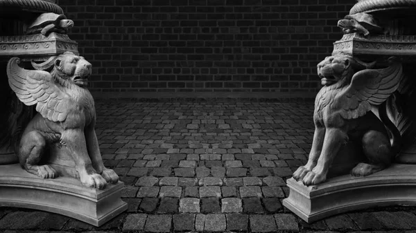 Two sculptures of winged lions stand on paving stones against a brick wall.