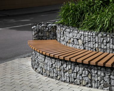 Metal ağın arkasındaki küçük taşlardan bir duvarın yanında eğri bir sıra düz tahta blok şeklinde bir park bankı..