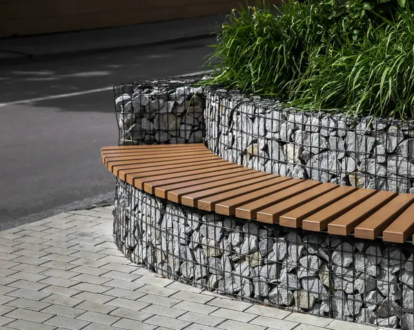 Metal ağın arkasındaki küçük taşlardan bir duvarın yanında eğri bir sıra düz tahta blok şeklinde bir park bankı..