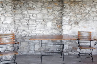 Sandalyeler ve arka planda taş bir duvarın karşısında bir masa.