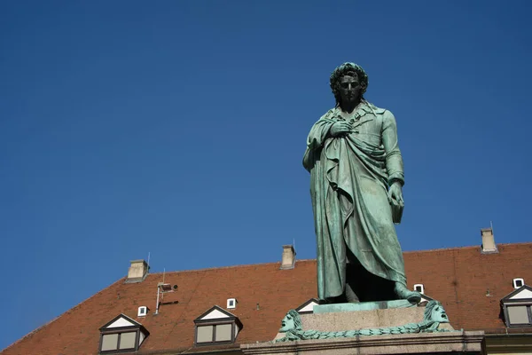 The larger-than-life bronze statue of Friedrich Schiller looks down at the viewer from its base on Shillerplatz. The tiled roof and some oriels of the Prince's Building can be seen behind the statue. A sunny day in Stuttgart without any clouds
