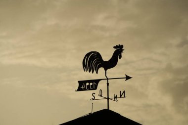 Gökyüzü akşam ışığı tarafından sarıya boyanmış ve bulutlarla dolu. Çatıdaki rüzgar horozu rüzgarın yönünü gösteriyor.