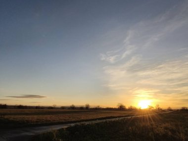 Uzak ormanın ağaçlarının arkasında güneş ufukta batıyor. Ön plandaki alan yolu hala biraz aydınlık ve güneşe giden yol gibi görünüyor.