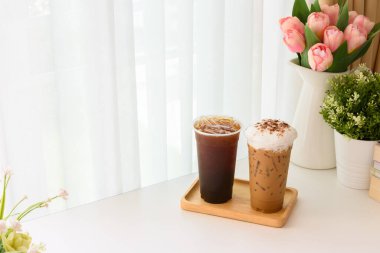 Ice Americano ve Ice Cappuccino kahvesi ve kapalı bir yerde beyaz perdeli, çiçeklerle süslenmiş ahşap bir tepsi..