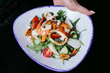 Karides, midye, kalamar, salatalık, kurdele, domates ve yeşil yapraklı taze deniz ürünleri salatası, mavi kenarlı beyaz bir kasede servis edilir..