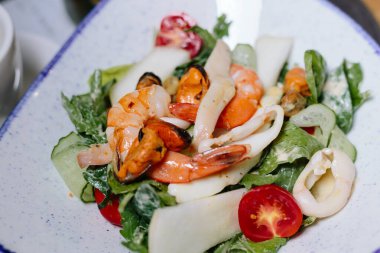 Karides, midye, kalamar, salatalık, kurdele, domates ve yeşil yapraklı taze deniz ürünleri salatası, mavi kenarlı beyaz bir kasede servis edilir..