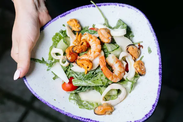 Karides, midye, kalamar, salatalık, kurdele, domates ve yeşil yapraklı taze deniz ürünleri salatası, mavi kenarlı beyaz bir kasede servis edilir..