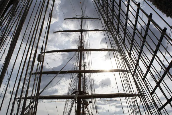 Bir yelkenli gemisinin teçhizatı ana direği ve merdiven kablolarını detaylı olarak gösteriyor ve bulutlu bir günde güneş içinden bakarken yelken açıyor.